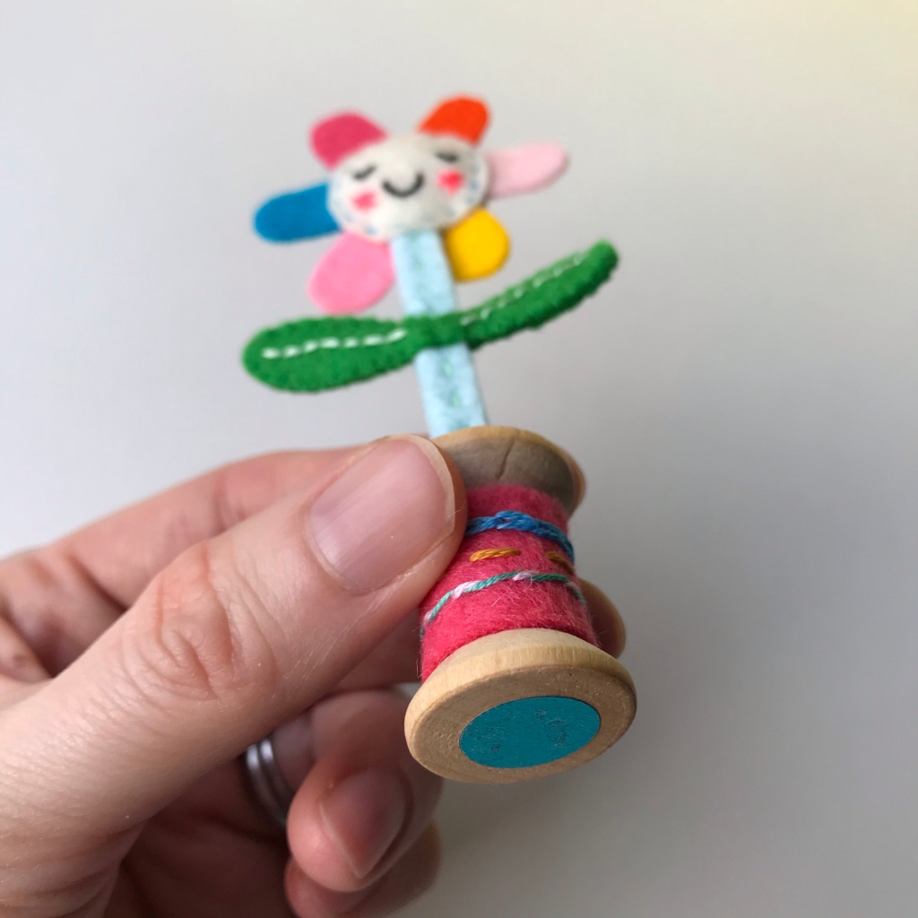 Hand holding a mini happy flower at an angle so you can see the bottom of the wooded bobbin with a shiny metallic blue sticker.